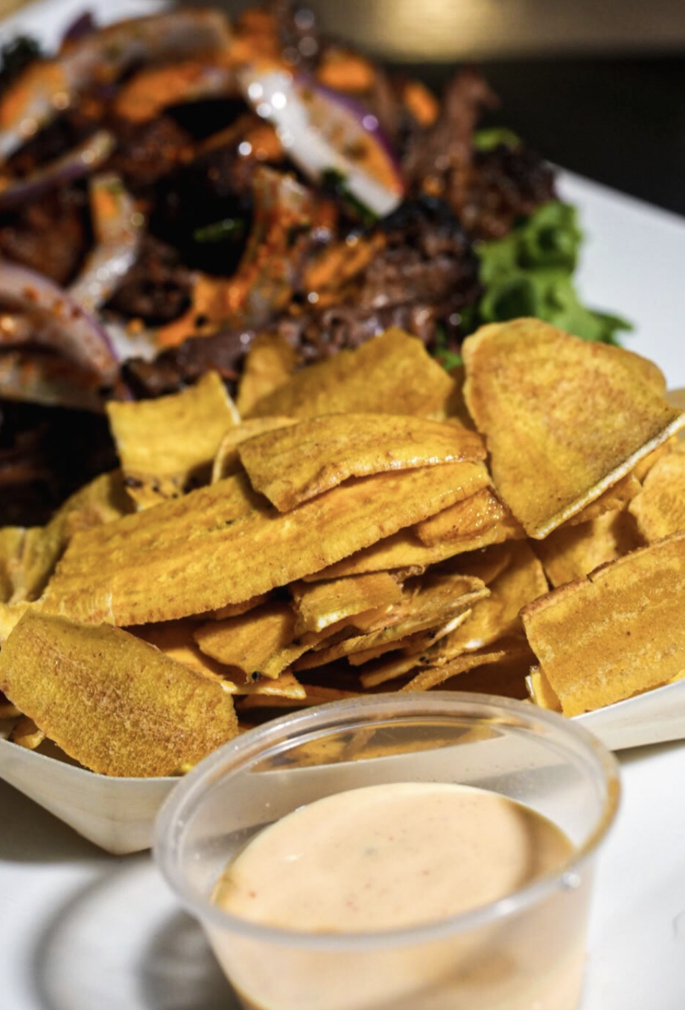 Crisp plantain chips with creamy dipping sauce and a blurred braised beef slider in the background