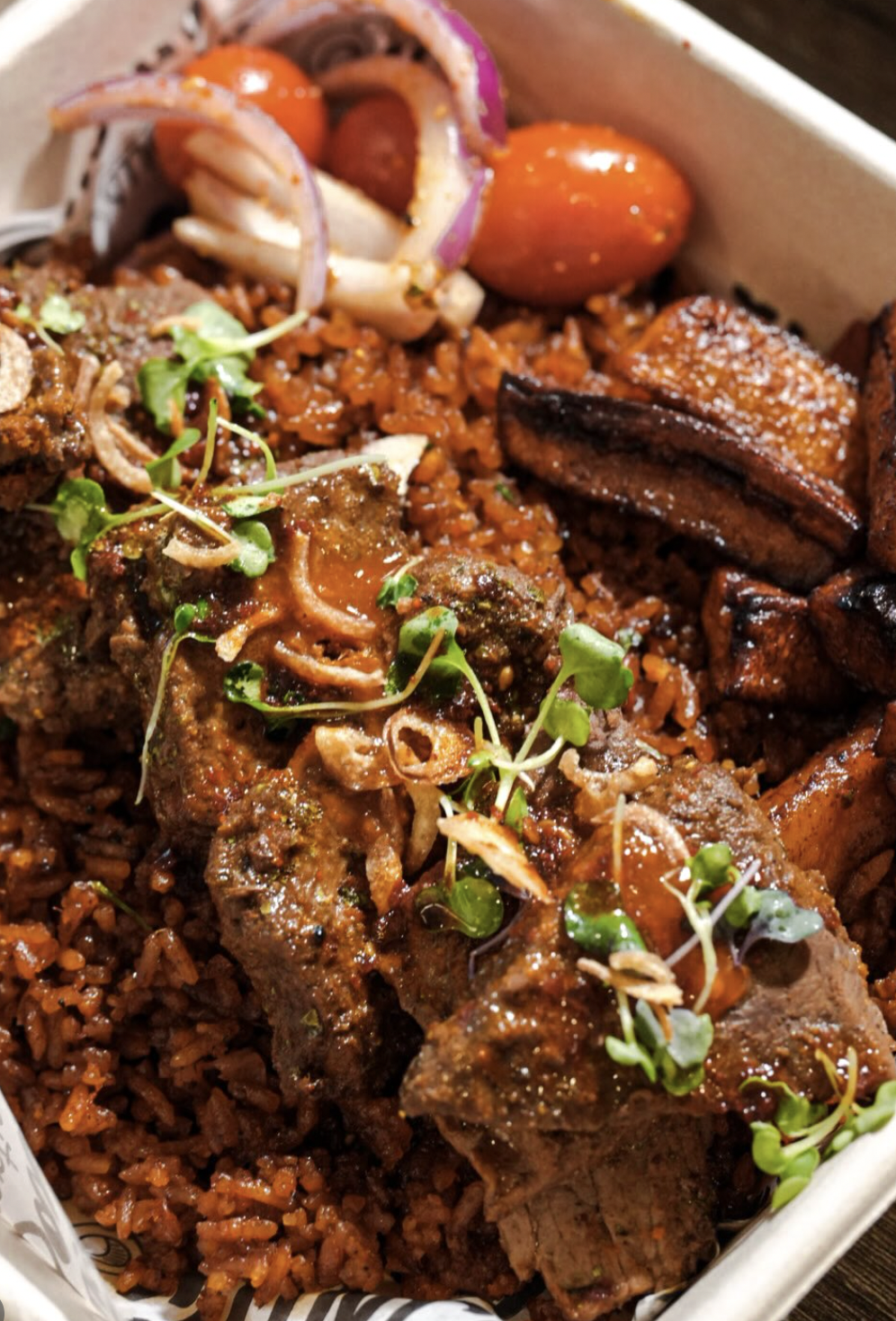 Jollof rice topped with lamb, plantains, tomatoes, and pickled onion