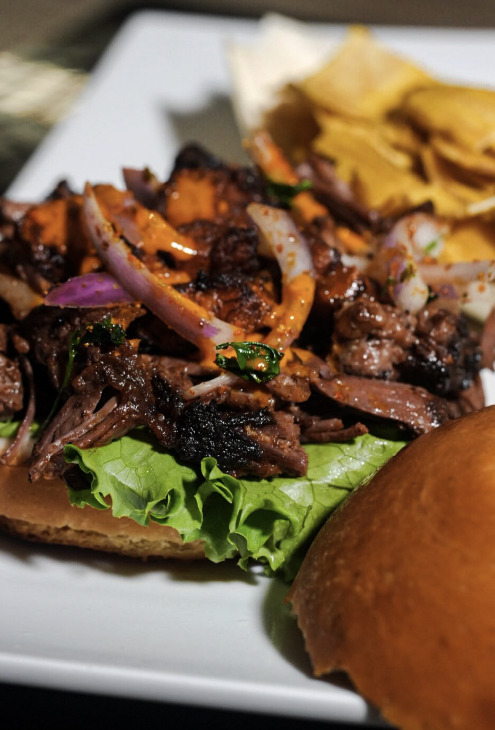Open-faced braised beef slider with lettuce, onions, and orange sauce beside plantain chips